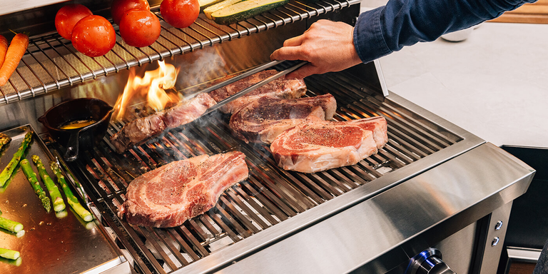 A close-up photograph captures thick ribeye steaks searing with a flare-up on a professional stainless steel grill, while asparagus and tomatoes cook alongside.