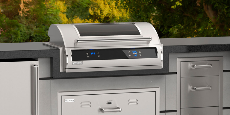 Modern stainless steel electric grill with digital temperature controls built into a sleek outdoor kitchen island, surrounded by greenery.