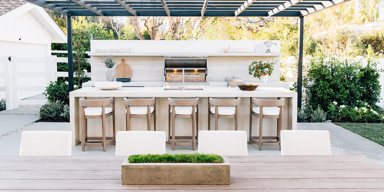 A straight-on view of the minimalist outdoor kitchen from kitchenlayout9.jpg, showing five wooden barstools lined up against a thick white stone island. Behind it, a matching white counter holds a stainless steel grill and a single floating shelf.