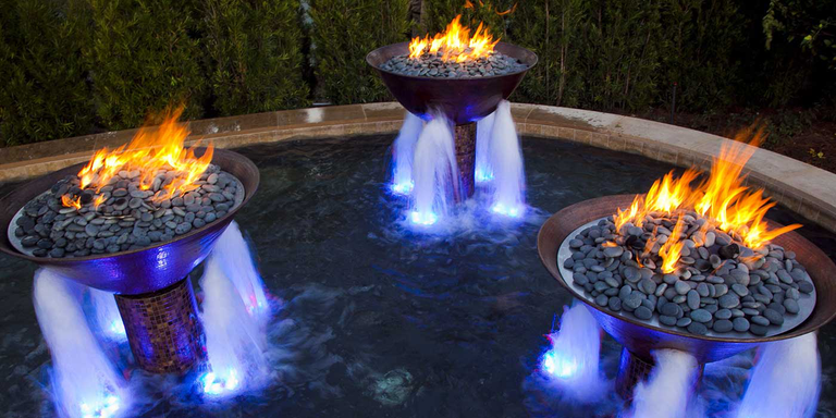 Three copper fire bowls with water pouring out of the sides installed in the center of a large, round pool.