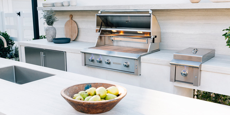Modern outdoor kitchen featuring a built-in stainless steel gas grill with open lid, side burner, and clean white countertops, with a bowl of green apples in the foreground and minimalist decor in the background.