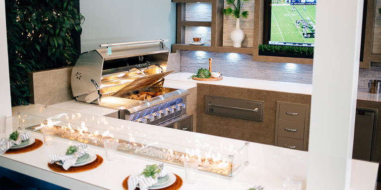 A luxury gallery kitchen featuring a high-end stainless steel grill with blue LED control knobs and an open hood revealing food inside. The space includes white quartz countertops, a long built-in glass-encased fire pit integrated into the dining peninsula, and a wall-mounted television displaying a football game. Modern stone-textured cabinetry, floating shelves with decorative vases, and formal place settings complete the sophisticated entertainment area.