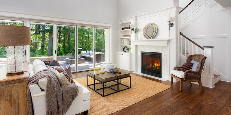 Elegant living room with white walls, hardwood floor, and large windows overlooking a green garden. Cozy fireplace, modern decor, and comfortable seating create a warm, inviting atmosphere.