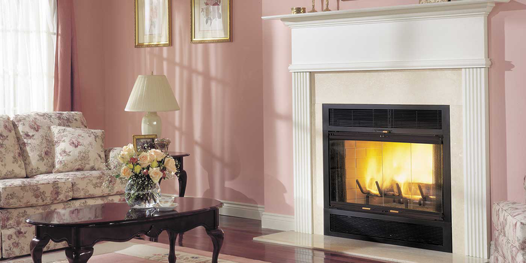 A French country-inspired living room with light pink walls, a white and pink floral print couch, an ornate wooden coffee table with a bouquet of flowers, a large, white hearth with a mantel and traditional, square wood burning fireplace.