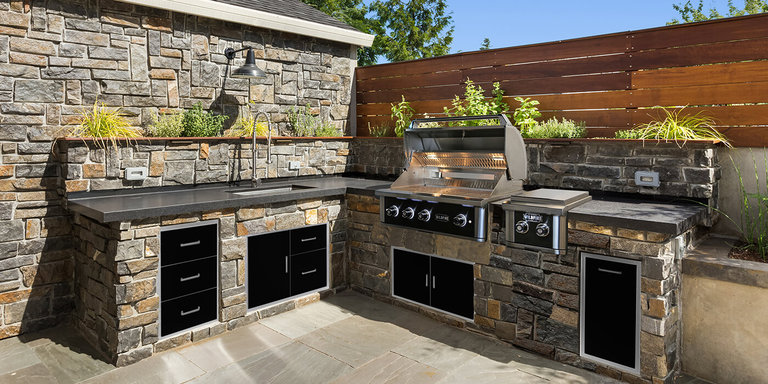 A rustic L-shaped outdoor kitchen built with multi-toned natural stone masonry and topped with dark gray countertops. The setup includes a large stainless steel grill with an open hood, a built-in side burner, and a deep sink with a high-arc faucet. Sleek black cabinetry and storage drawers are integrated into the stonework, while a horizontal wood slat fence and an herb garden planter ledge create a private, modern backdrop.
