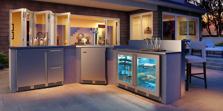 A sleek, contemporary outdoor bar and kitchen area at dusk, featuring stainless steel appliances with bright blue interior lighting. The L-shaped counter includes a double-tap beer tower and glass-front refrigerators stocked with beverages.