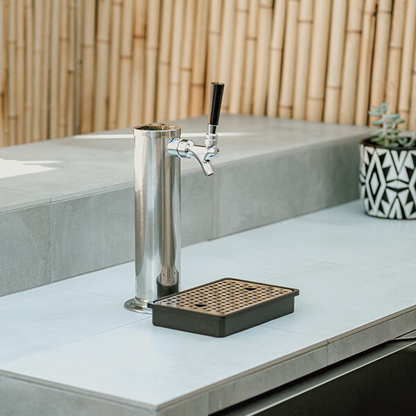 A stainless steel single-tap draft tower with a black handle is mounted on a light grey tiled countertop, featuring a black drip tray directly beneath the spout. The modern outdoor setup is positioned against a natural bamboo fence background, with a small potted succulent visible on the right.