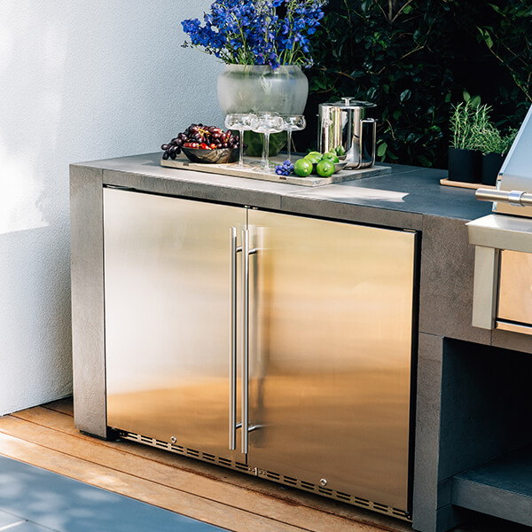 Stainless steel outdoor beverage center with double doors, topped with fruit, glassware, and a drink setup on a modern patio counter.