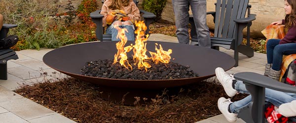 A family of five gathered around a large round fire pit on a stone patio on a cool evening. A man stands smiling while two teenagers and two younger children sit in dark gray Adirondack chairs, some wrapped in cozy plaid blankets. The patio is set against a natural sandstone retaining wall with autumn-colored plantings, and a large window reflects the dusky sky behind them.