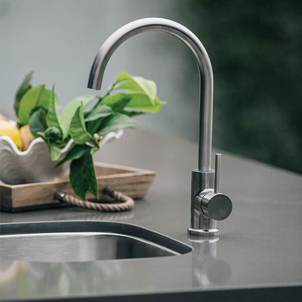 A high-angle, close-up shot of a modern, brushed stainless steel gooseneck faucet mounted on a grey countertop next to a small undermount sink. In the soft-focus background, a wooden tray holds a white scalloped bowl filled with lemons and leafy green branches.