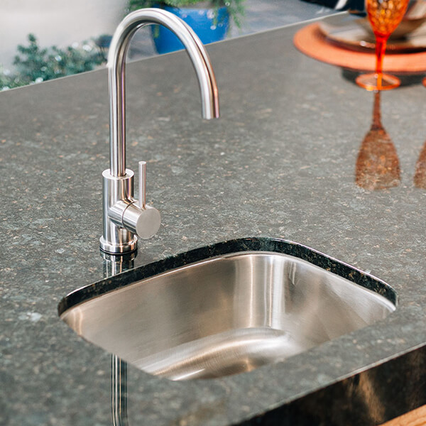 A high-angle view of a sleek, brushed stainless steel gooseneck faucet and a small undermount sink set into a dark, speckled granite countertop. In the soft-focus background, an orange glass and a blue planter add a pop of color to the modern outdoor bar area.